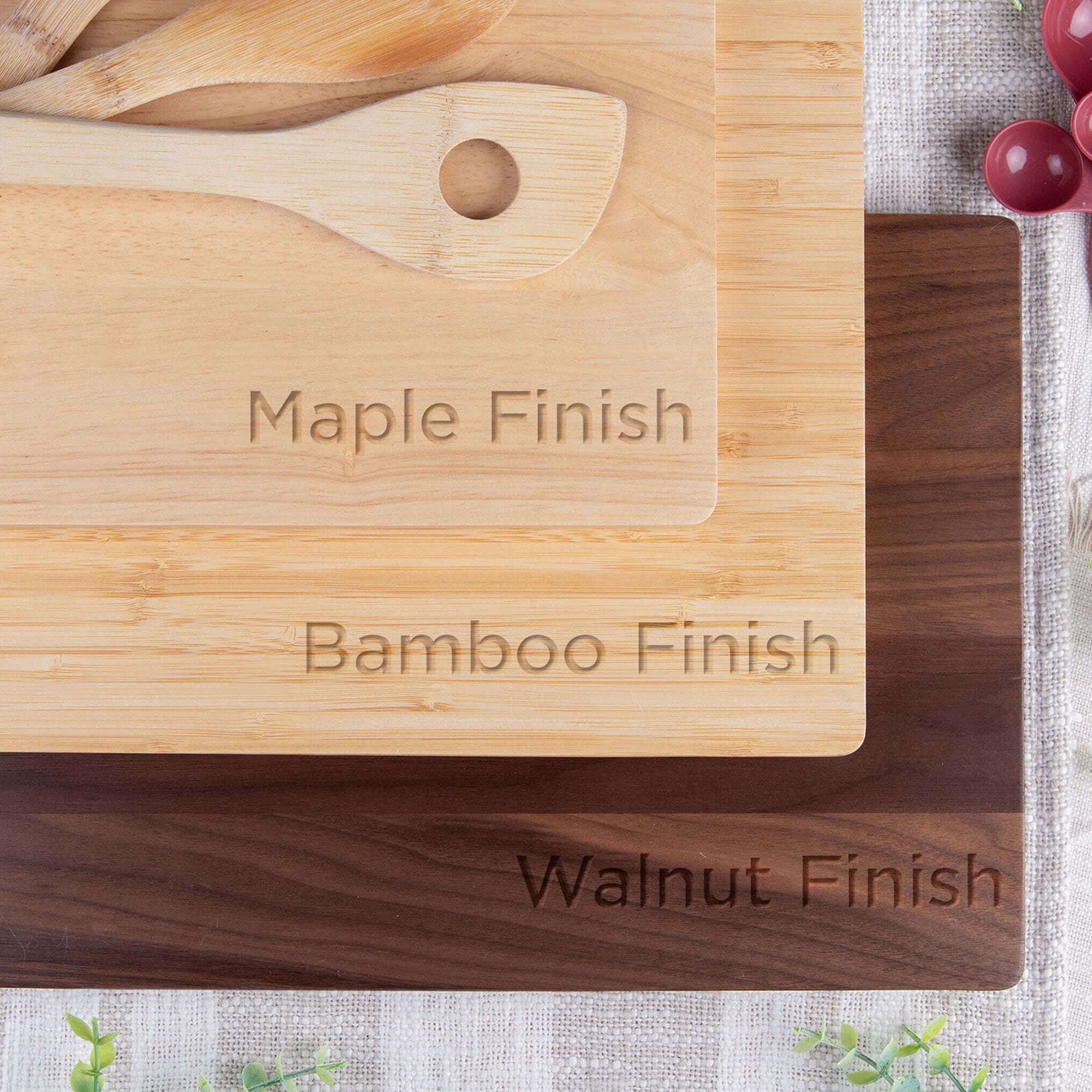 Stacked engraved cutting boards displaying maple, bamboo, and walnut finishes with wooden spoons and measuring cups, showcasing material and color options for custom engraved cutting boards.