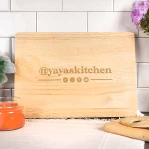 Maple wood cutting board engraved with the text ‘@yayaskitchen’ and social media icons, styled in a bright kitchen setting with a white tile backsplash, orange candle, and purple flowers.