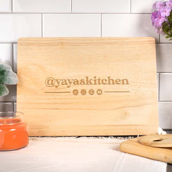 Maple wood cutting board engraved with the text ‘@yayaskitchen’ and social media icons, styled in a bright kitchen setting with a white tile backsplash, orange candle, and purple flowers.