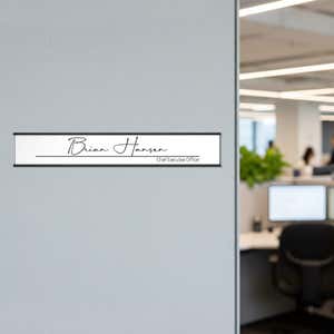 A white name plate with a black aluminum holder on an office wall