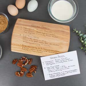 Maple cutting board engraved with a handwritten glazed pecans recipe, displayed on a dark surface with ingredients including eggs, brown sugar, milk, pecans, and a matching handwritten recipe card.