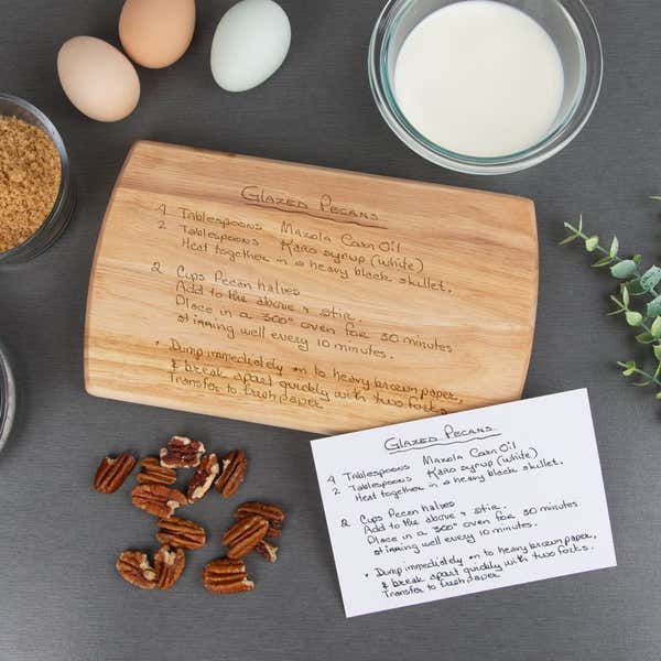 Maple cutting board engraved with a handwritten glazed pecans recipe, displayed on a dark surface with ingredients including eggs, brown sugar, milk, pecans, and a matching handwritten recipe card.